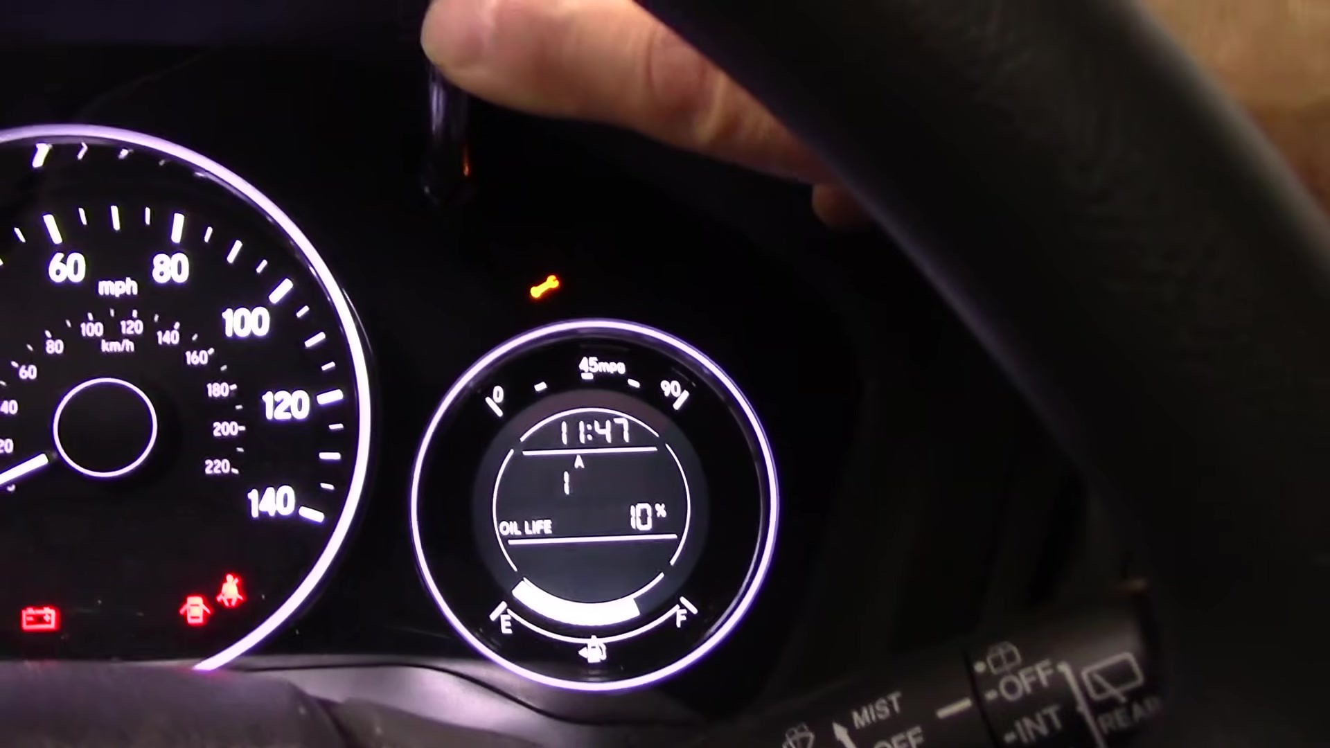 Honda HR-V Mk2 cluster showing Oil Life 10% and the amber maintenance wrench, with a finger pressing the select/reset button beside the display.