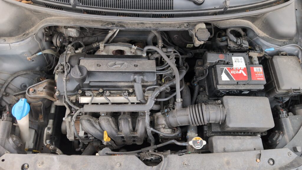 Engine-bay view of the Hyundai i20 Mk2 (GB) 12V battery with cover and terminal clamps.