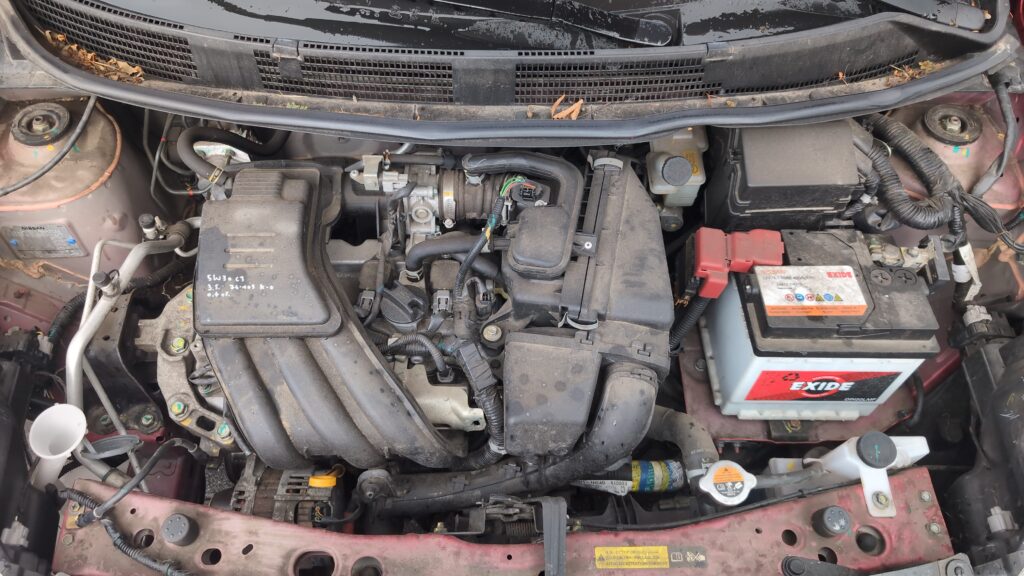 Engine-bay view of the Nissan Micra K13 12V battery with cover and terminal clamps.