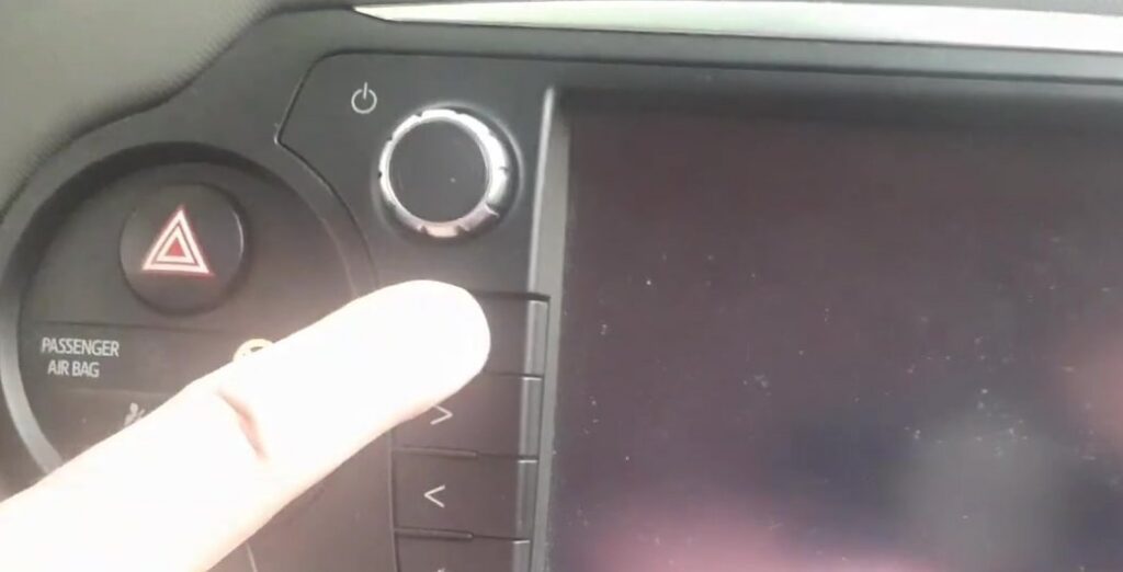 Finger reaching for the Toyota Touch 2 audio power knob (top-left of the screen, marked with the power symbol) on a Toyota Yaris XP130, the control you press and hold to open the Service Menu