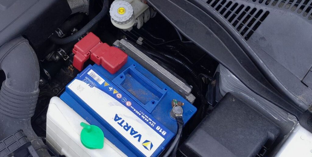 12V battery in the engine bay of a Suzuki Swift with its positive terminal cover